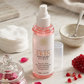 Skincare toner bottle with pink liquid on a wooden surface with flowers and a towel.