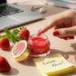 Hand applying luxova red lip balm to a small jar with strawberries and a laptop in the background