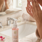 Person applying toner to face with a bottle of toner on a bathroom counter.